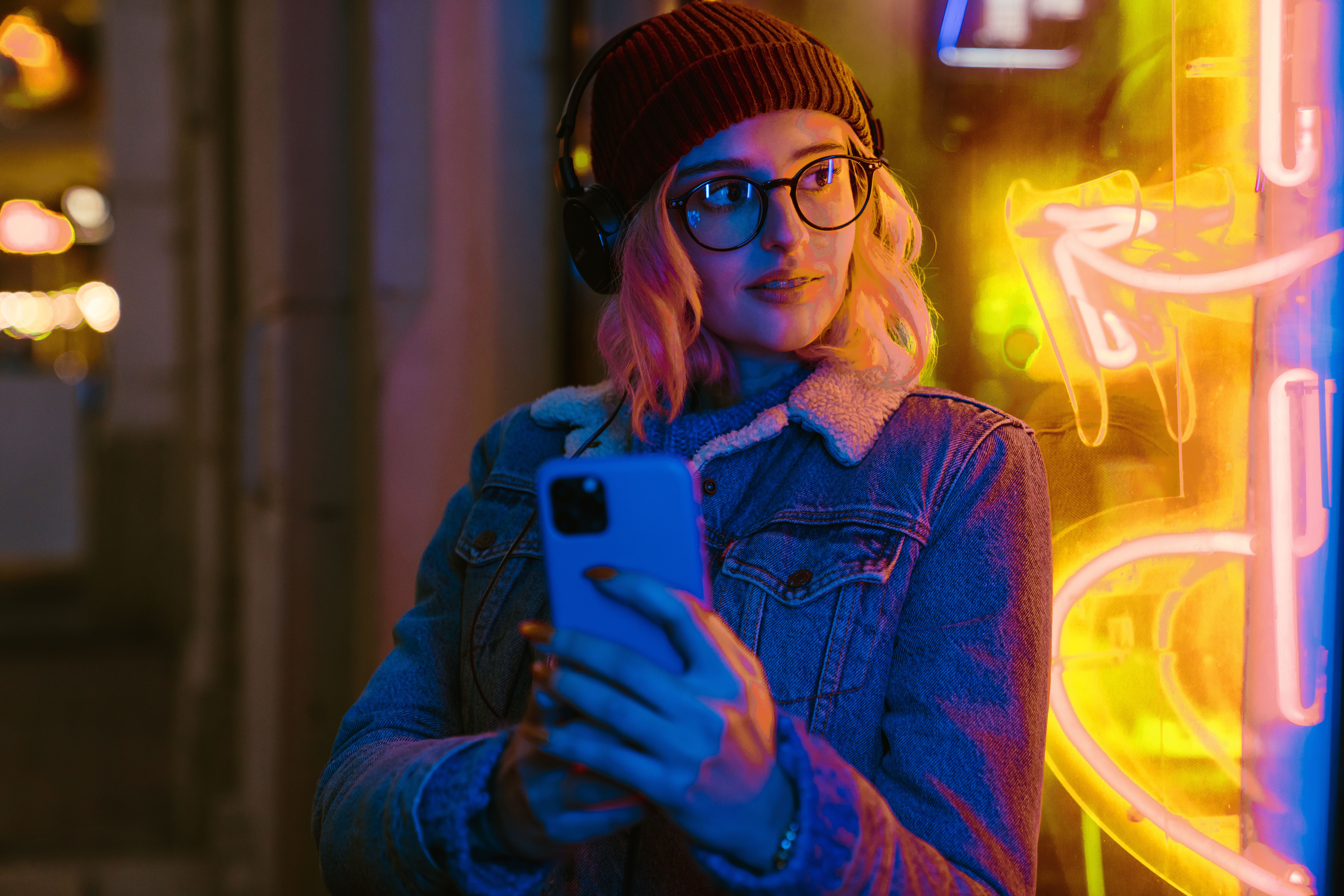 Young woman listening music and using cellphone at city street Woman holding phone in front of neon sign